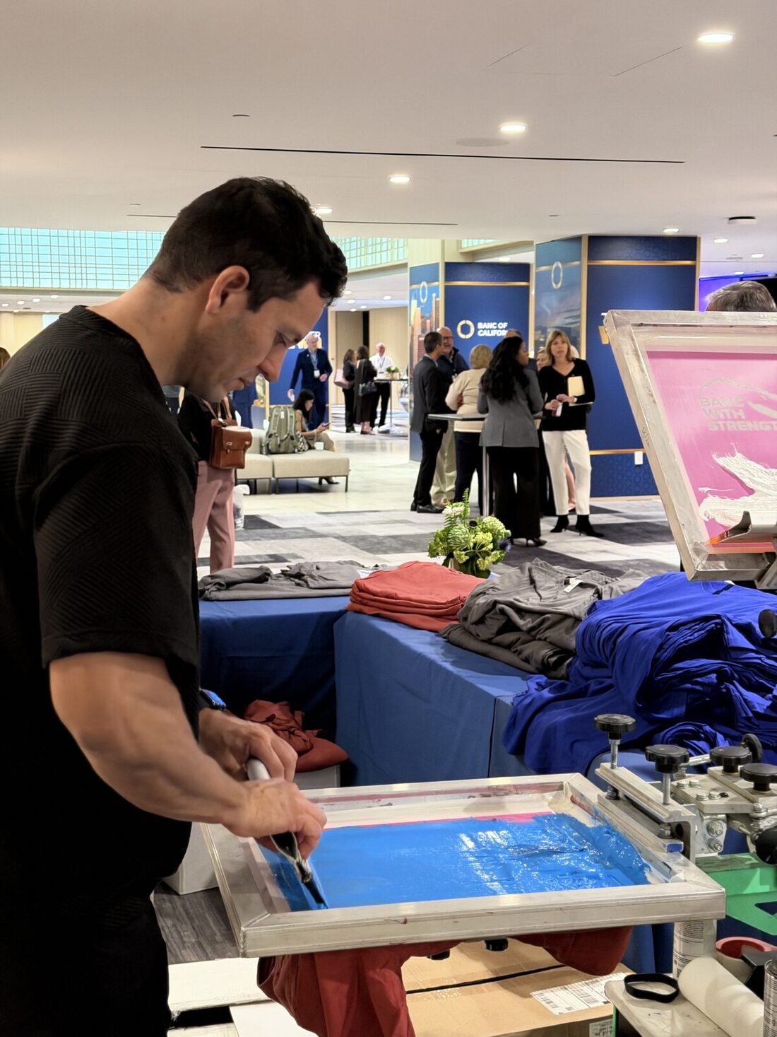 A Merch Troop printer pulling a squeegee across a screen on a press at an indoor New York corporate event, with folded shirts and guests in the background